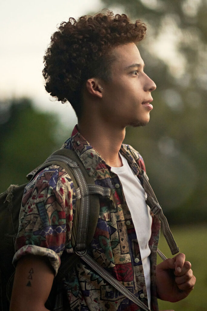 A man outdoors with a thoughtful expression, carrying a backpack and looking into the distance.