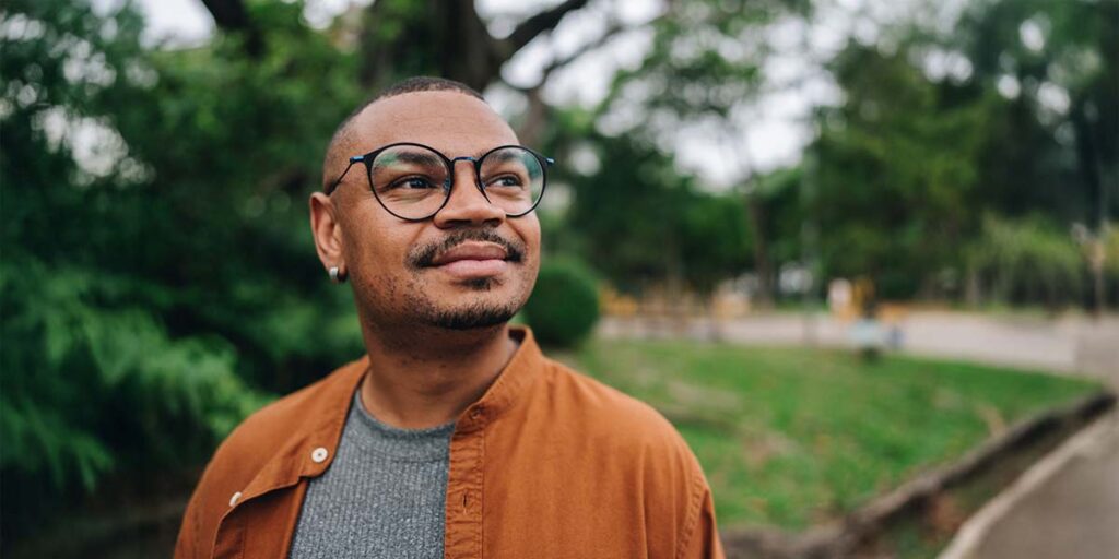 A man with glasses and a warm smile, standing outside in a green park.